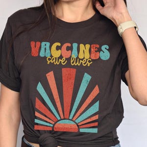 May include: A dark grey t-shirt with a retro-style graphic that says "Vaccines save lives" in colorful letters. The graphic also features a stylized sunrise with red, orange, and blue stripes.
