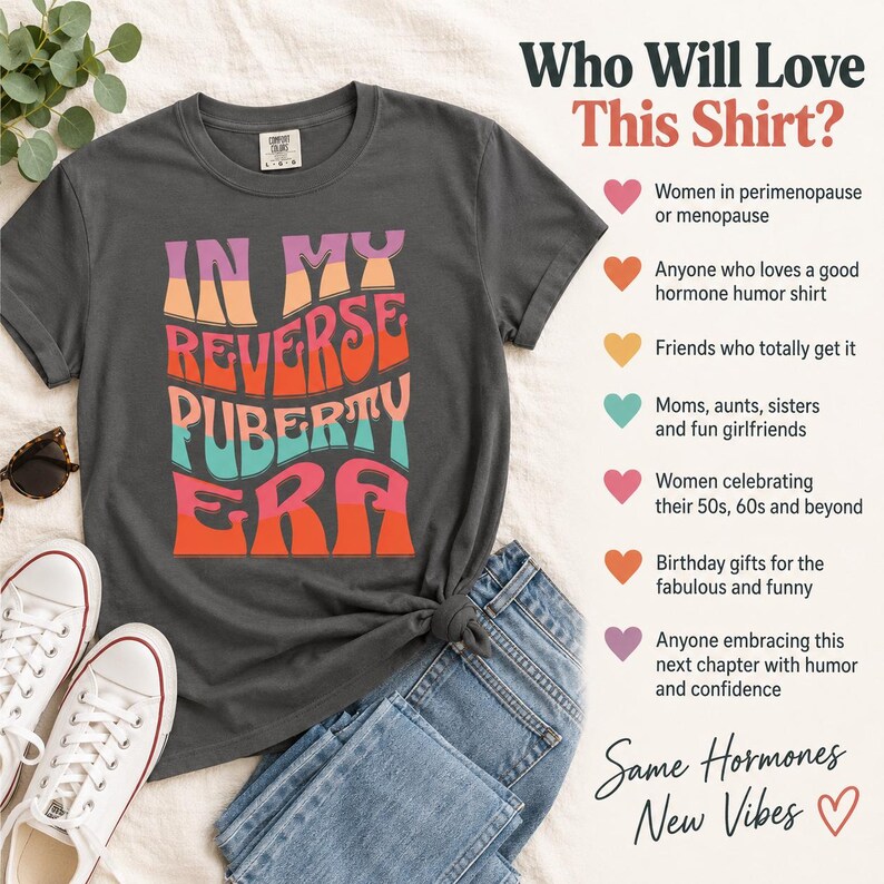 Op de afbeelding: Een antracietgrijs t-shirt met de tekst "IN MY REVERSE PUBERTY ERA" in een golvend, veelkleurig lettertype in roze, oranje en turquoise. Het t-shirt wordt getoond naast een blauwe spijkerbroek, witte sneakers en een zonnebril op een textiel ondergrond.