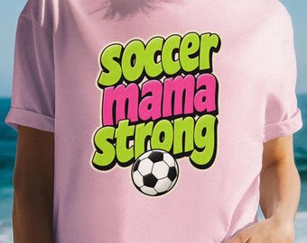Op de afbeelding: Een lichtroze t-shirt met de tekst "soccer mama strong" in een vet, gelaagd lettertype. De woorden "soccer" en "strong" zijn limoengroen, terwijl "mama" felroze is. Een zwart-witte voetbalafbeelding bevindt zich onderaan de tekst.