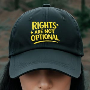May include: Black baseball cap featuring the embroidered phrase "RIGHTS ARE NOT OPTIONAL" in yellow. The cap has a curved peak and a button on top. The text is centered on the front of the cap.