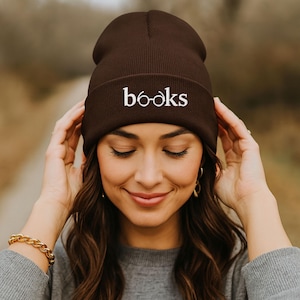 May include: A dark brown beanie with the word "BOOKS" embroidered in white. The beanie is made of a soft, ribbed knit material. The hat is being worn by a person.