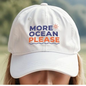 May include: A white baseball cap featuring the embroidered text MORE OCEAN PLEASE in blue and orange. The design includes a small orange sun icon above the text and a blue wavy line underneath, with a button on top and stitched eyelets.
