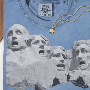 May include: A light blue Comfort Colors t-shirt featuring a graphic of Mount Rushmore with surprised expressions. A gold star pendant hangs from a gold chain. The shirt is size XL-TG-EG.