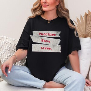 May include: Black t-shirt with the text "Vaccines Save Lives" in red letters. The text is arranged on three horizontal, torn-paper-style banners. The shirt is a casual, oversized fit.