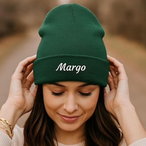 May include: A dark forest green knit beanie featuring a folded cuff. The word Margo is embroidered in white cursive script across the center of the cuff. The hat has a ribbed texture and a rounded crown, providing a simple and functional head covering.