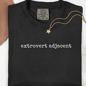 May include: Black t-shirt with the phrase "extrovert adjacent" in white lettering. A gold-colored necklace with a star pendant is draped over the shirt. The shirt has a Comfort Colors tag.