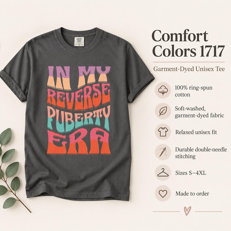 Op de afbeelding: Een antracietgrijs t-shirt met korte mouwen en de tekst IN MY REVERSE PUBERTY ERA in golvende, veelkleurige letters in roze, oranje en turquoise. Het shirt is gemaakt van 100% ringgesponnen katoen met dubbele stiksels en een relaxte pasvorm.