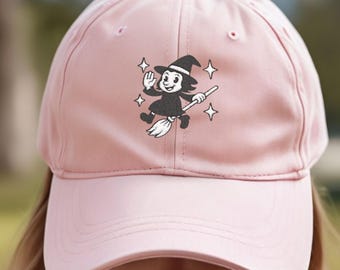 May include: A light pink baseball cap featuring a black and white cartoon witch graphic. The witch is riding a broom and surrounded by stars. The cap has a curved brim and a button on top.