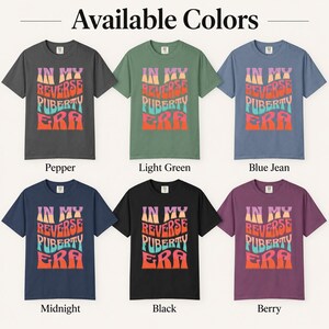 Op de afbeelding: Zes t-shirts met korte mouwen in de kleuren pepper, light green, blue jean, midnight, black en berry. Elk shirt is voorzien van de tekst IN MY REVERSE PUBERTY ERA in een golvende, meerkleurige grafische print met tinten paars, oranje en blauwgroen.
