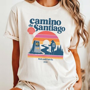May include: A cream-colored t-shirt featuring a graphic of a hiker, a winding path, a milestone marker with a scallop shell, and a sunset. The text reads "camino de Santiago", "Rob and Fidelia", and "2026" in dark blue lettering on the front.