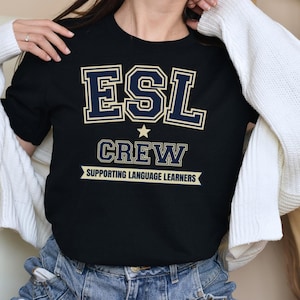 May include: Black crew neck t-shirt with the text "ESL CREW SUPPORTING LANGUAGE LEARNERS" in navy blue and cream-colored lettering. The shirt has a star graphic above the word "CREW".