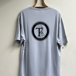 May include: A light blue t-shirt with a black circle logo on the back. The logo has the word "TAWAKAL" written around the outside and a stylized "T" in the center.