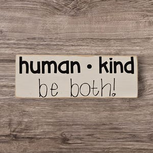 May include: A rectangular, off-white wooden sign with the words "human • kind be both!" in black lettering. The sign is set against a rustic, wooden background.