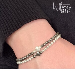 May include: Two bracelets with silver and black beads, each with a silver heart charm. The bracelets are on a person's wrist.