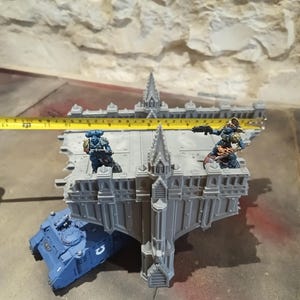 Op de afbeelding: Een grijze miniatuurgebouw in gotische stijl met een blauwe tank en twee miniatuurfiguren. Het gebouw is ongeveer 30 cm lang. De figuren dragen gevechtsuitrusting. De scène speelt zich af op een grijs oppervlak met een lichtgekleurde stenen muur op de achtergrond.