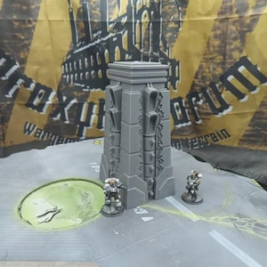 Può includere: Un pezzo di terreno grigio rettangolare con un design industriale dettagliato. Due figure in miniatura sono in piedi nelle vicinanze su una superficie grigia con un elemento circolare verde. Lo sfondo presenta uno striscione giallo e nero con un'illustrazione di fabbrica.