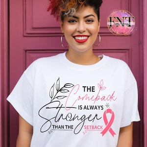 May include: White t-shirt with the text "The Comeback is Always Stronger Than The Setback" in black and pink. The design includes a pink ribbon and floral elements. The model is smiling.