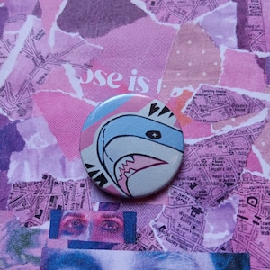 Puede incluir: Un botón redondo con un diseño de tiburón de dibujos animados. El tiburón tiene un cuerpo azul claro, dientes blancos y detalles rosas. El botón está sobre un fondo de papel rosa rasgado con imágenes de mapas y ojos.