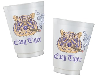 May include: Two translucent white plastic cups, each featuring a graphic illustration of a tiger wearing purple lightning bolt sunglasses and smoking a cigar. The text "Easy Tiger" is printed in purple below the tiger illustration on both cups.