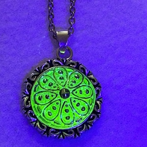 May include: A round pendant necklace featuring a vibrant, glowing neon green center with a symmetrical floral-like pattern of teardrop shapes and dots, encased in an ornate, dark metallic filigree frame, suspended from a matching metal chain.