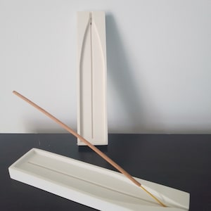 Peut inclure: Deux porte-encens en céramique blanche. Le plus grand est un long rectangle étroit avec une rainure au centre. Le plus petit est un rectangle plus court et plus large avec une rainure au centre. Un bâton d'encens brun est posé sur le dessus du plus grand porte-encens.