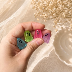 Puede incluir: Cuatro pequeñas figuras translúcidas con forma de fantasma en azul, verde, rosa y morado. Cada fantasma presenta un rostro negro impreso sencillo con dos ojos circulares y una boca ovalada, elaborado con un material liso y brillante.