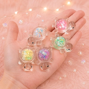 May include: Four clear resin turtle figurines with hollow shells filled with colourful confetti shapes including stars, hearts, and circles. Each turtle has a smooth, transparent body, black dot eyes, and a small red painted mouth line.