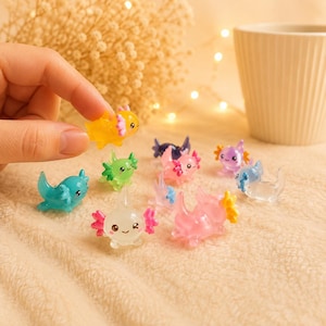 Pode incluir: Uma coleção de pequenas figuras de axolote translúcidas em várias cores, incluindo amarelo, verde, azul, rosa e roxo. Cada criatura em miniatura apresenta um acabamento liso e brilhante, traços faciais arredondados e brânquias externas.