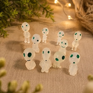 Puede incluir: Una colección de diez pequeñas figuras blancas translúcidas con cabezas lisas y redondeadas y ojos circulares verdes. Cada figura presenta una expresión facial única, incluyendo puntos simples o pequeñas líneas para representar la boca.