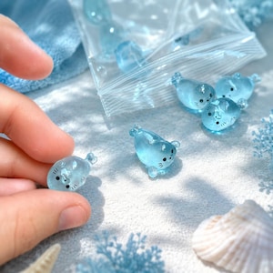 May include: Small, translucent light blue glass seal figurines with black painted eyes and whiskers. Several of these miniature animal charms are scattered on a surface, with some contained inside a clear plastic resealable storage bag.