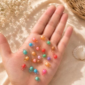 May include: A collection of small, miniature turtle figurines in various translucent and solid colours including pink, blue, green, orange, and purple. Each tiny turtle features a detailed shell pattern and is scattered across the palm of a hand.