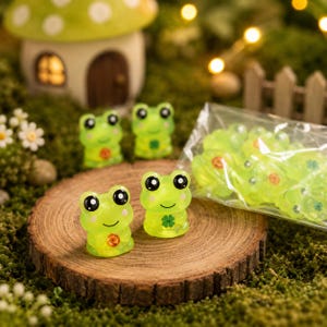 Puede incluir: Una colección de pequeñas figuras de ranas de resina en color verde lima translúcido. Cada rana tiene grandes ojos negros, mejillas rosadas y una boca sonriente. Algunas ranas muestran una pequeña flor naranja o un trébol de cuatro hojas.