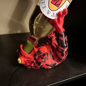 May include: A red and black frog-shaped bottle holder with a yellow eye, holding a bottle of white wine with a label that says "Tequila Lime" and a red and yellow logo.