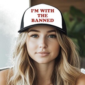 May include: A black and white trucker hat with the text "I'M WITH THE BANNED" in red. The hat has a white front panel and a black mesh back. The person in the image is wearing a gray tank top.