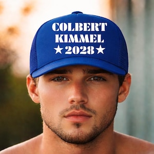 May include: A blue trucker hat with the text "COLBERT KIMMEL 2028" in white. The hat has a mesh back and a curved brim. The hat is worn by a person.