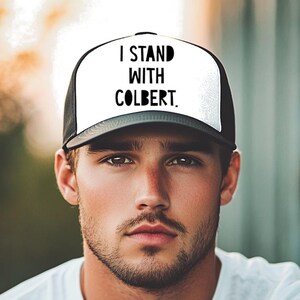 May include: Black and white trucker hat with the text "I STAND WITH COLBERT" in bold, black letters. The hat has a white front panel and a black mesh back. The person in the image is wearing a white t-shirt.