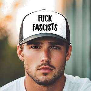 May include: A black and white trucker hat with the text "FUCK FASCISTS" in bold, black letters. The hat has a white front panel and a black mesh back. The person in the image is wearing a white t-shirt.