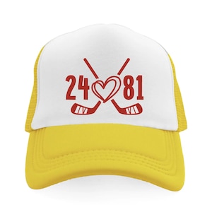 May include: A yellow and white trucker hat with a red graphic. The graphic features the numbers 24 and 81, crossed hockey sticks, and a heart. The hat has a mesh back and a curved brim.