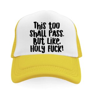 May include: A yellow and white trucker hat with the text "This too SHALL PASS. BUT Like. HOLY FUCK!" in black. The hat has a yellow brim and mesh backing, and a white front panel.