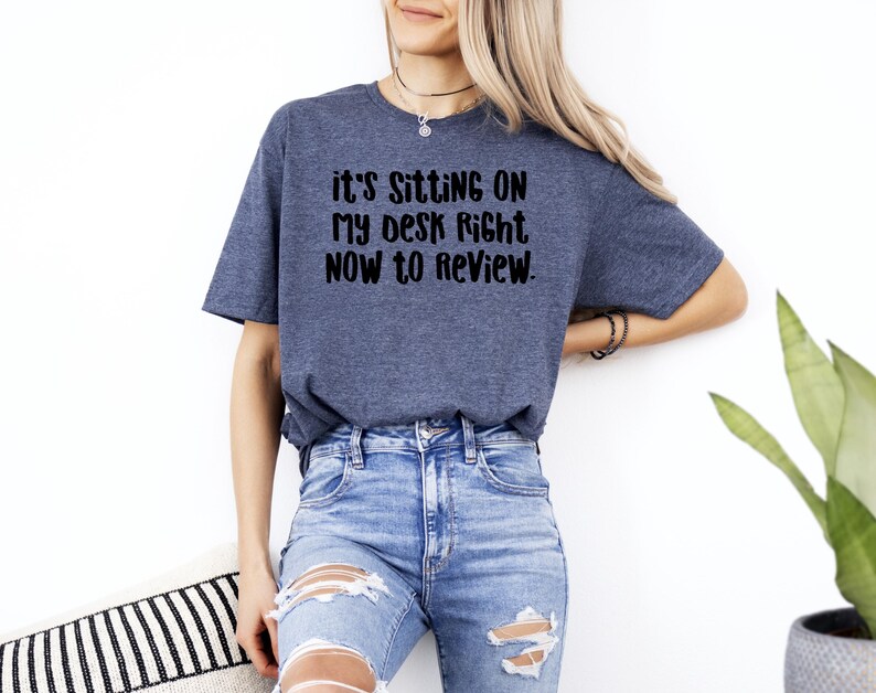 May include: A blue t-shirt with the text "it's sitting on my desk right now to review" in black. The shirt is paired with blue jeans. The model is wearing a necklace and bracelets.