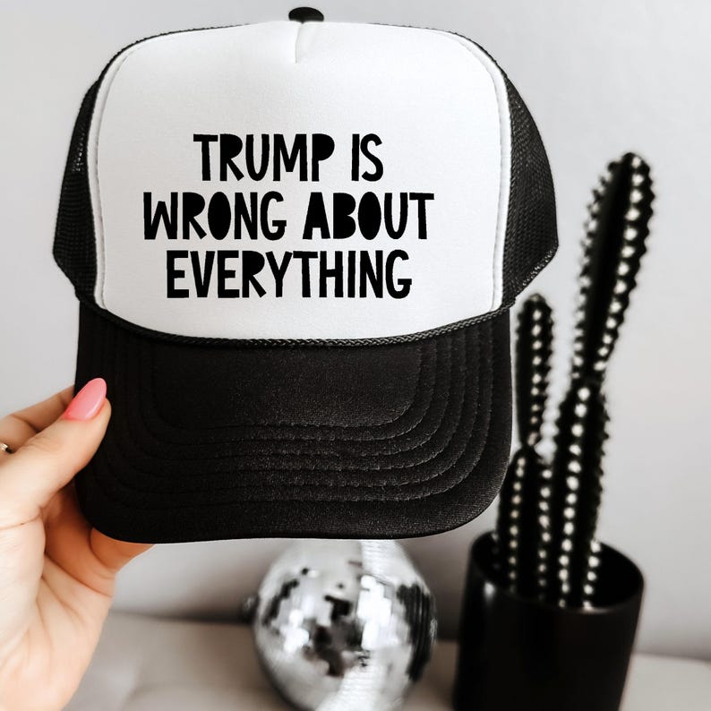 May include: Black and white trucker hat with the text "TRUMP IS WRONG ABOUT EVERYTHING" in bold, black letters. The hat has a white front panel and a black brim and mesh back. A hand is holding the hat.