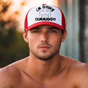 May include: A red and white trucker hat featuring a white front panel with a black graphic of a torso and the text I'M GOING COMMANDO in black lettering. The hat has a red brim and a red mesh back panel with an adjustable snapback closure.