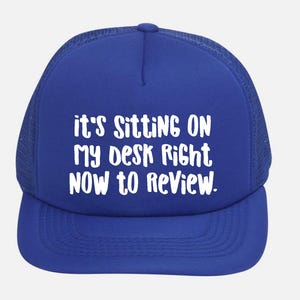 May include: Royal blue trucker hat with white text that reads "it's sitting on my desk right now to review." The hat has a mesh back and a curved brim. A casual accessory.