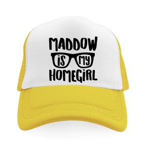 May include: A yellow and white trucker hat with the text "MADDOW IS MY HOMEGIRL" in black. The hat has a white front panel and a yellow brim and mesh back.