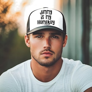 May include: A black and white trucker hat with the text "JIMMY IS MY HOMEBOY" in black lettering. The hat has a white front panel and a black brim and mesh back. The person is wearing a white t-shirt.