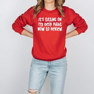 May include: A red sweatshirt with the text "it's sitting on my desk right now to review" in white. The long-sleeved crew neck sweatshirt is worn with light blue jeans.