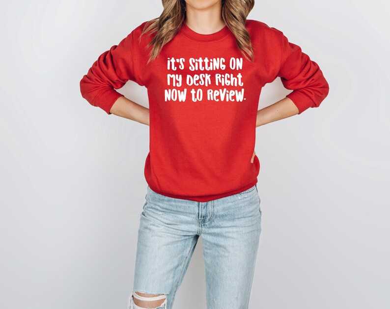 May include: A red sweatshirt with the text "it's sitting on my desk right now to review" in white. The long-sleeved crew neck sweatshirt is worn with light blue jeans.