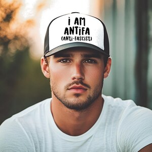 May include: A black and white trucker hat with the text "I AM ANTIFA (ANTI-FASCIST)" in bold, black letters. The hat has a white front panel and a black mesh back. The person in the image is wearing a white t-shirt.