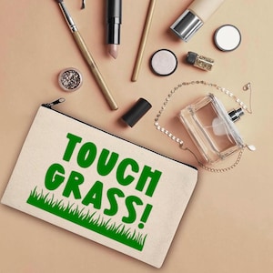 May include: A beige canvas cosmetic bag with a black zipper and the text "TOUCH GRASS!" in green. Below the text is a green grass graphic. Makeup brushes, lipstick, and perfume are scattered around the bag.