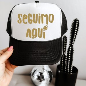 May include: A black and white trucker hat with the text "SEGUIMO AQUI" in gold lettering and a football graphic. The hat has a mesh back and a curved brim. The hat is being held by a person.